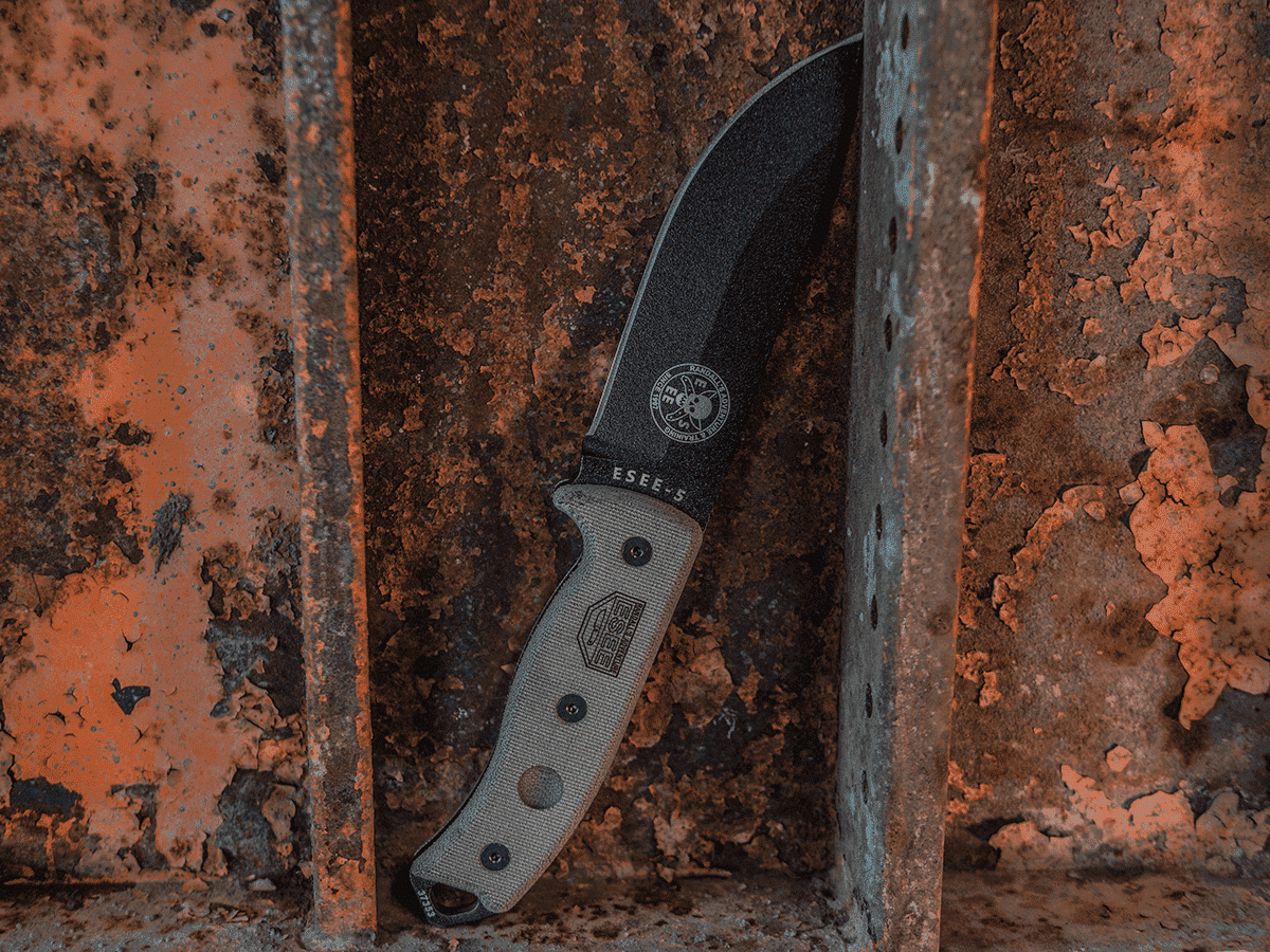 Das ESEE-5 Survivalmesser mit dicker, schwarzer Klinge und ergonomischem Micarta-Griff, auf rustikalem Untergrund präsentiert.